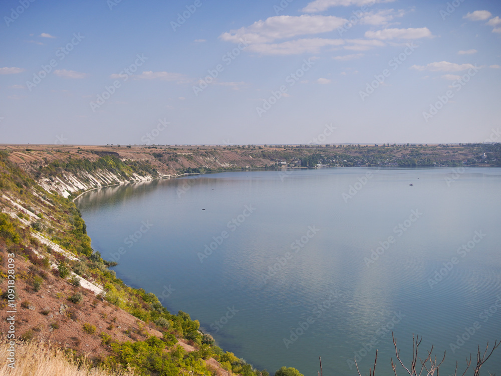 Obraz premium A calm Dniester River stretches under a clear sky. The river curves gently, bordered by a steep, earthy bank covered in sparse vegetation. A small village is visible in the distance across the water.
