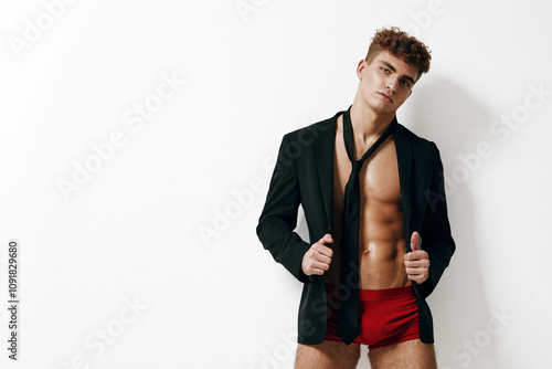 Confident young man with curly hair posing in an unbuttoned blazer, wearing red shorts, exuding charm and allure against a minimalist white background. photo