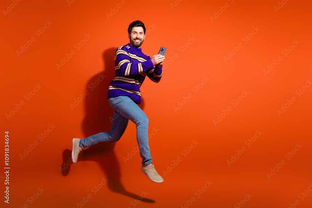 Fototapeta premium Full body photo of attractive young man running hold gadget dressed stylish striped purple clothes isolated on orange color background
