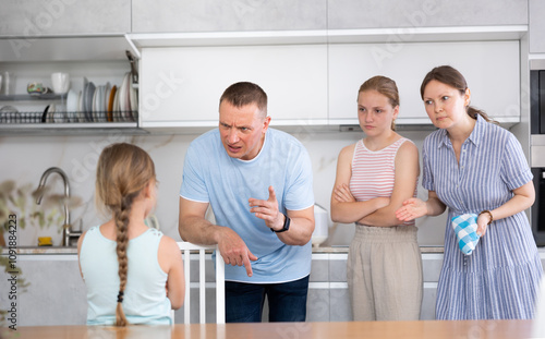 Preteen girl with long pigtail standing in home kitchen, listening to disapproving words of father, mother and older sister. Concept of tension and disagreement arising between generations photo