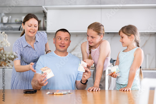 Man is sitting near kitchen table and counting bills. He gives his smiling wife and daughter money for ladie' nice little things, trinkets photo