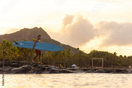 A man is walking on a jetty in Waikiki with a surfboard in his hand. The sky is cloudy and the sun is rising.