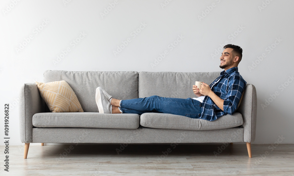 © Prostock-studio - Rest Concept. Happy Arab guy drinking coffee sitting on comfortable couch at home in living room. Cheerful casual man relaxing on sofa, enjoying weekend free time or break from work, full body length