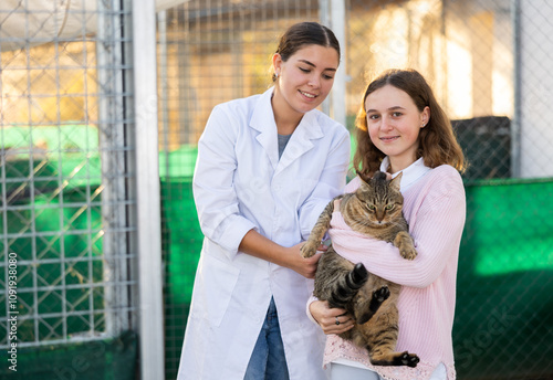 Cheerful cute tween girl standing near outdoor enclosure for homeless animals in shelter, holding big gray tabby tomcat. Pet adoption and children social responsibility concept .. photo