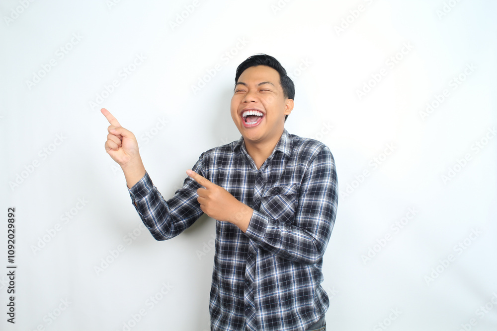 Fototapeta premium carefree asian man pointing hand with laughing expression showing joy and playful emotion wearing casual plaid shirt isolated on white background
