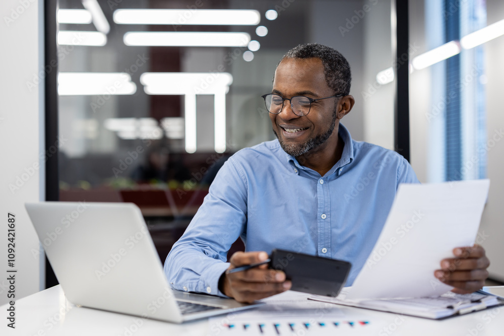 Mature African American businessman smiling while reviewing documents in modern office setting with laptop and smartphone. Professional environment emphasizes productivity, achievement, success. Mature African American businessman smiling while reviewing documents in modern office setting with laptop and smartphone. Professional environment emphasizes productivity, achievement, success.