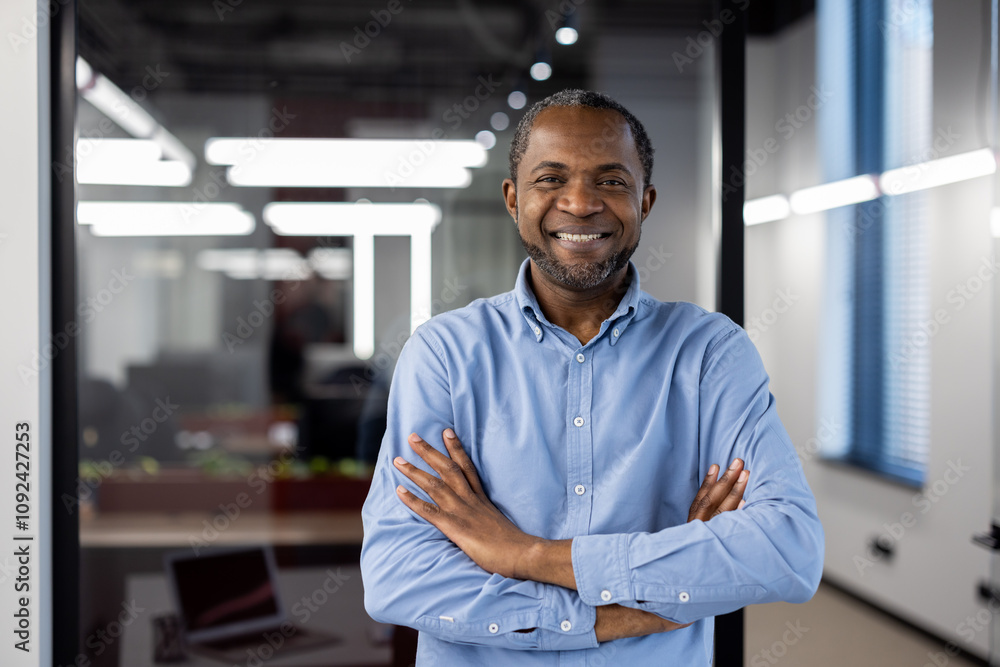 Obraz premium Confident African American businessman standing in modern office. Man wearing blue shirt, smiling with arms crossed. Corporate environment with bright lighting and glass partitions.