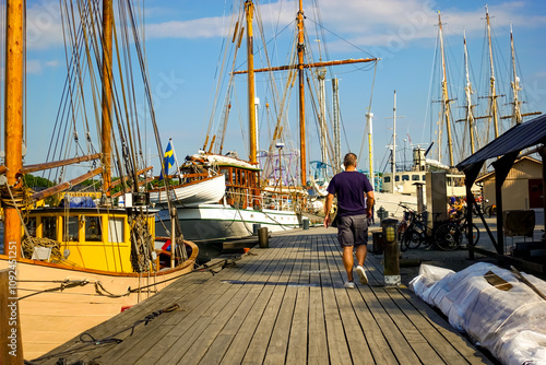 Adult man walking on a wooden plank pathway by old sailboats in a historical Swedish marina on a summer afternoon with masts in the background against blue sky photo