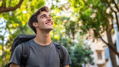 A young man with a backpack smiles while looking up in a green, outdoor setting.