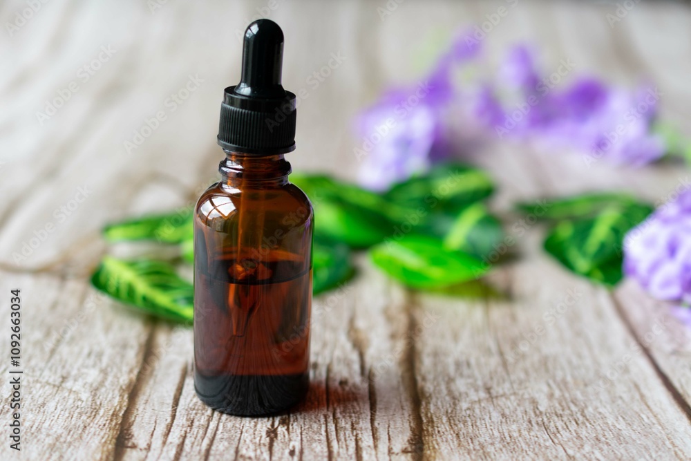 Amber vial of essential oil with a dropper, on a wooden surface, with purple flowers and leaves in the background Amber vial of essential oil with a dropper, on a wooden surface, with purple flowers and leaves in the background