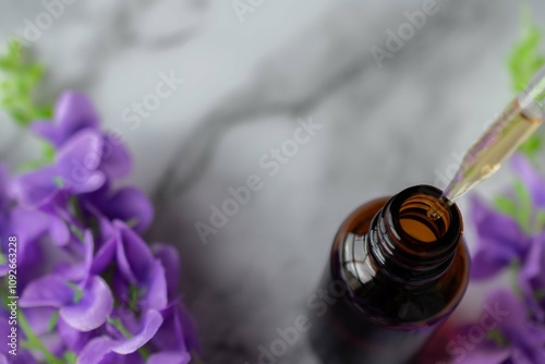 Close-up of a dropper taking some essential oil from an amber vial, on a marble surface with some flowers around it.