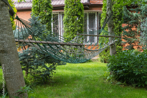 Holiday atmosphere. bright sunny day. Wicker hammock stretched between trees, against blurred background of evergreen garden and country house. Selective focus.