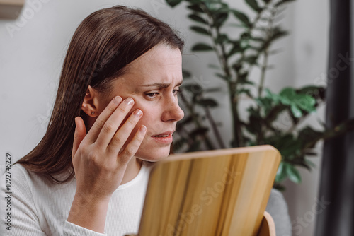 Unhappy young caucasian woman looking in mirror, allergy presenting allergic reaction from cosmetic on face. Anti wrinkle aging concept. Beauty, health care from acne problem, dry skin, treatment