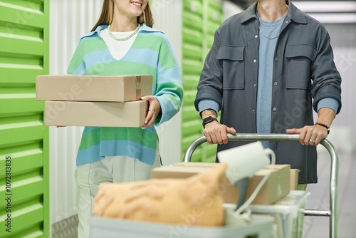 Cropped shot of smiling woman carrying pile of cardboard boxes walking next to boyfriend helping with moving out, while pushing loaded dolly cart in hallway of self storage warehouse photo