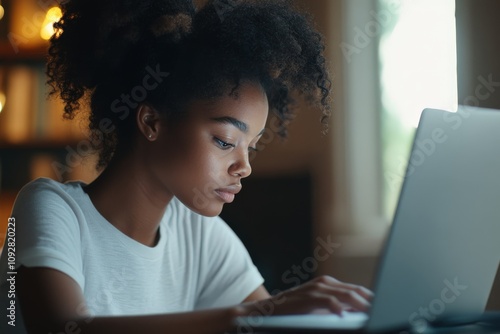 A young woman with curly hair deeply focused on working at her laptop in a warmly lit room, representing concentration, dedication, and modern technology.