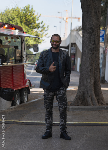 A man in a black jacket and camouflage pants gives a thumbs up, ok while standing on a quiet urban street beside a red food truck. The casual outdoor setting conveys a relaxed and friendly atmosphere