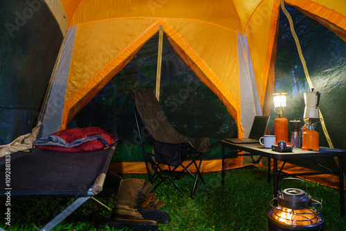 Camping tent, folding bed inside the tent at night in the forest, gas lantern and coffee set, notebook on a wooden camping table, and outdoor ideas.