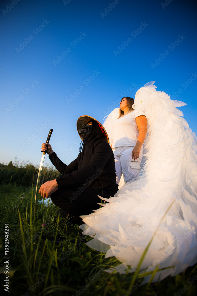 Naklejka premium Handsome samurai and angel against blue sky. A masked samurai kneels on a grassy field holding a katana next to a man with large white wings resembling an angel under a clear blue sky.