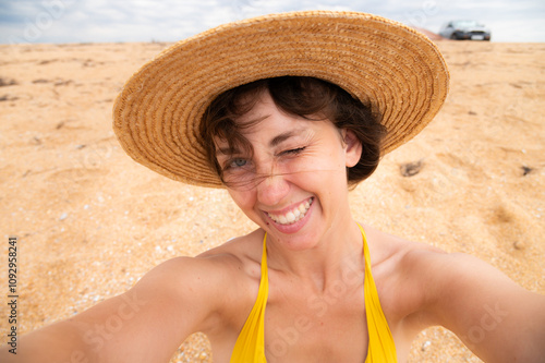 Close up shot of good looking female tourist enjoys free time outdoor near ocean on beach, looks at camera during leisure on sunny summer day, poses for selfie. Happy smiling tourist in tropics #1092958241