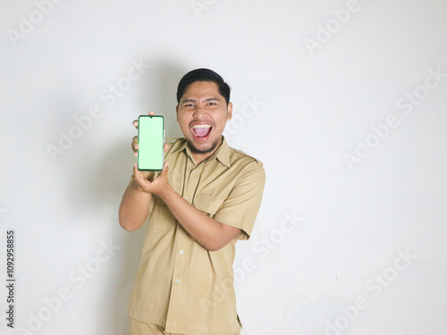 An Asian male Indonesian civil servant wearing a brown uniform, stands and shouted, screaming while holding a cellphone while showing a blank or greenscreen. Indonesian civil servants, ASN photo