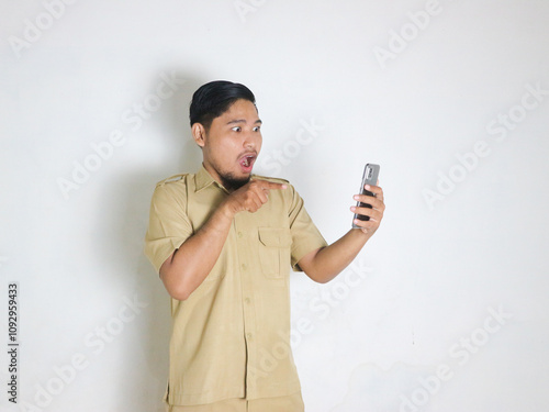 An Asian male Indonesian civil servant wearing a brown uniform who is usually called a keki shows a gesture of surprise and shock while looking at his mobile phone. Indonesian civil servants, ASN photo