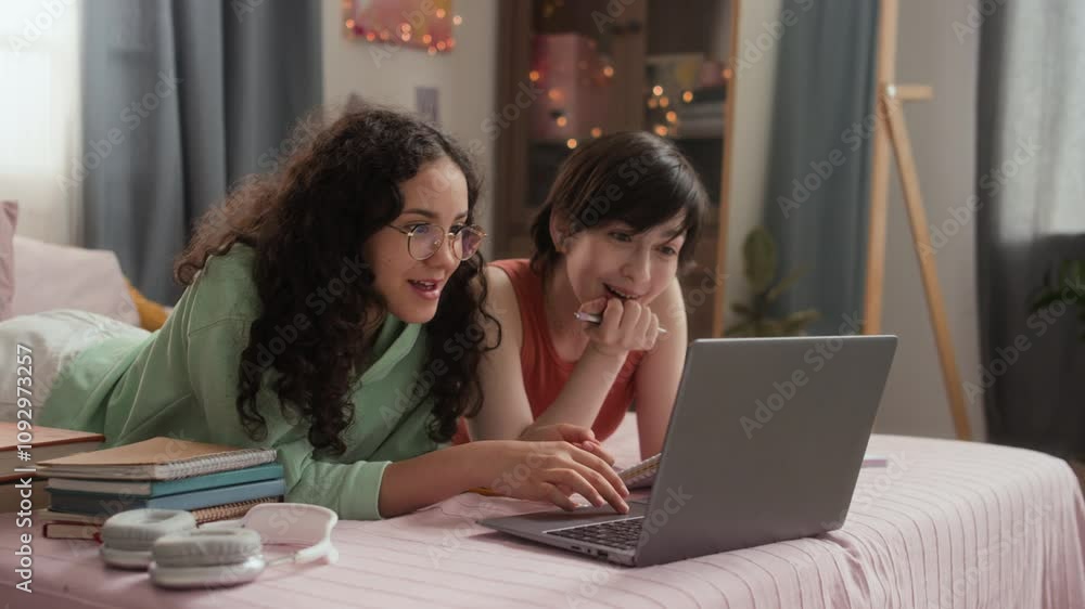 Two teenage girls lying on bed, looking at laptop screen with smiles on their faces, enjoying studying time together in cozy room Two teenage girls lying on bed, looking at laptop screen with smiles on their faces, enjoying studying time together in cozy room