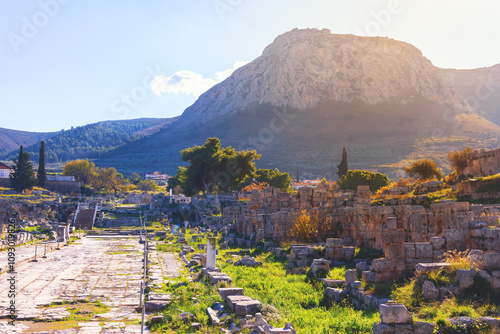 View of the Corinth Archaeological Park with ancient ruins, a stone path, and Acrocorinth mountain in the background, under bright sunlight. Corinth, Greece photo