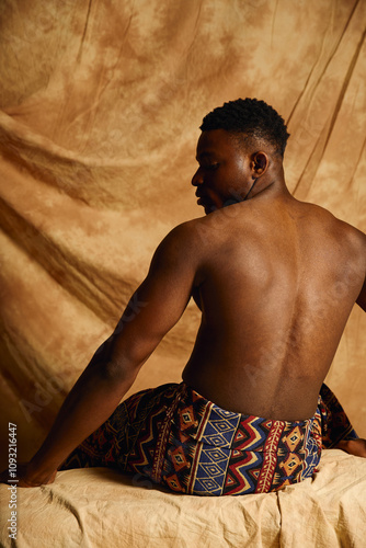 A young handsome african american man poses topless, exuding confidence and charm. He is seated on a textured surface, dressed in a patterned wrap, with a neutral backdrop enhancing his features. photo