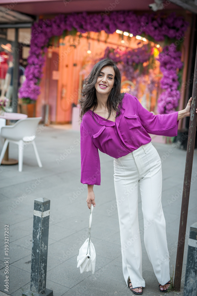 Young beautiful brunette woman in a pink jacket and white pants posing on the street. Portrait of a beautiful young woman in a purple blouse and white pants posing in an outdoor cafe on the street