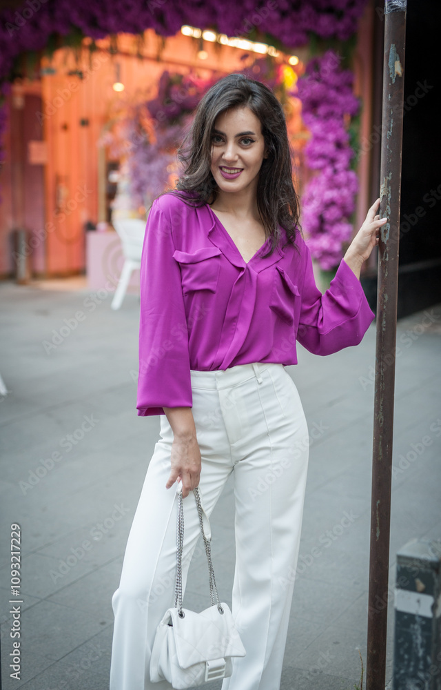 Young beautiful brunette woman in a pink jacket and white pants posing on the street. Portrait of a beautiful young woman in a purple blouse and white pants posing in an outdoor cafe on the street