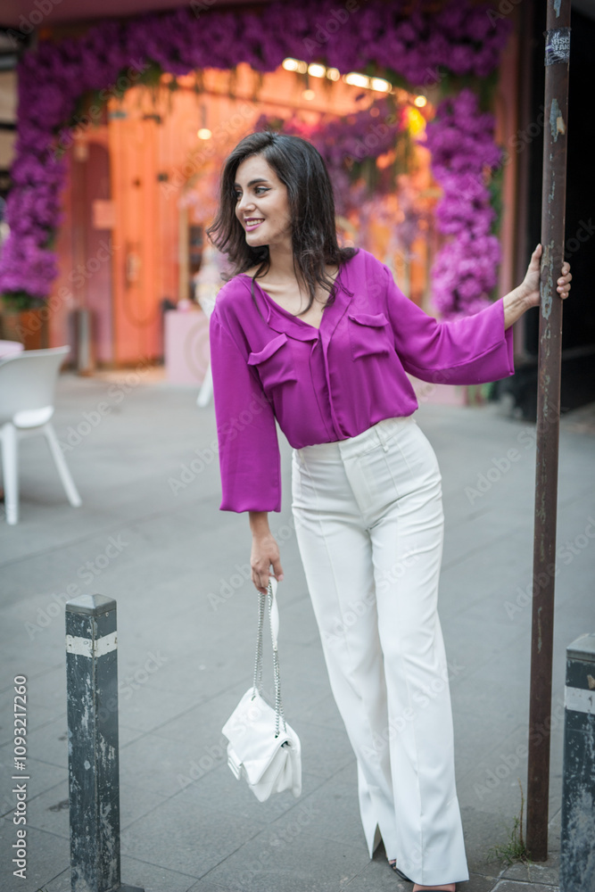 Young beautiful brunette woman in a pink jacket and white pants posing on the street. Portrait of a beautiful young woman in a purple blouse and white pants posing in an outdoor cafe on the street