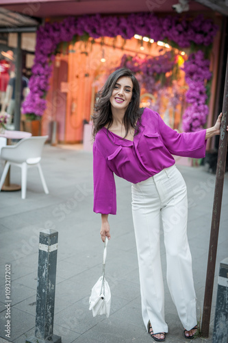 Obraz na płótnie Young beautiful brunette woman in a pink jacket and white pants posing on the street. Portrait of a beautiful young woman in a purple blouse and white pants posing in an outdoor cafe on the street w salonie