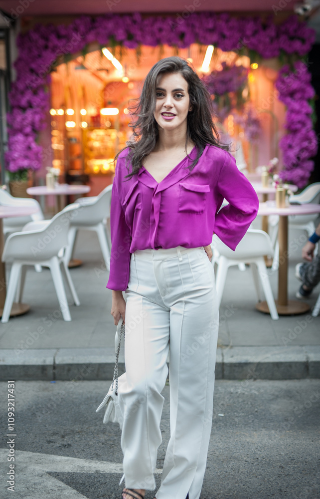 Young beautiful brunette woman in a pink jacket and white pants posing on the street. Portrait of a beautiful young woman in a purple blouse and white pants posing in an outdoor cafe on the street