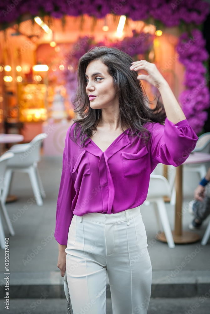 Young beautiful brunette woman in a pink jacket and white pants posing on the street. Portrait of a beautiful young woman in a purple blouse and white pants posing in an outdoor cafe on the street