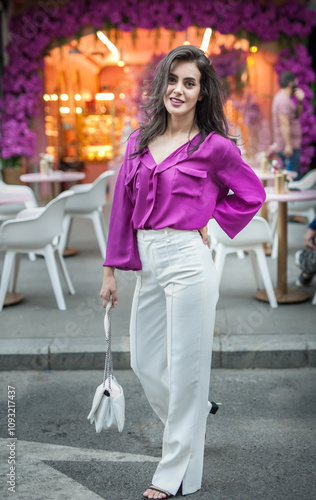 Young beautiful brunette woman in a pink jacket and white pants posing on the street. Portrait of a beautiful young woman in a purple blouse and white pants posing in an outdoor cafe on the street