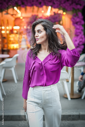 Obraz na płótnie Young beautiful brunette woman in a pink jacket and white pants posing on the street. Portrait of a beautiful young woman in a purple blouse and white pants posing in an outdoor cafe on the street w salonie