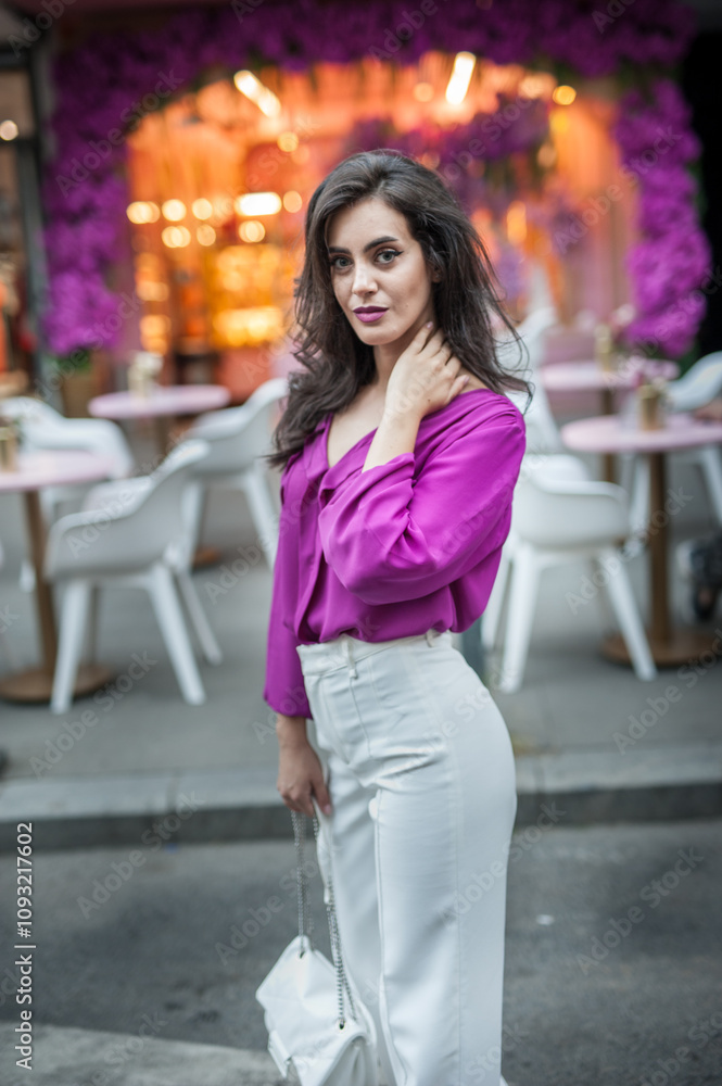 Young beautiful brunette woman in a pink jacket and white pants posing on the street. Portrait of a beautiful young woman in a purple blouse and white pants posing in an outdoor cafe on the street