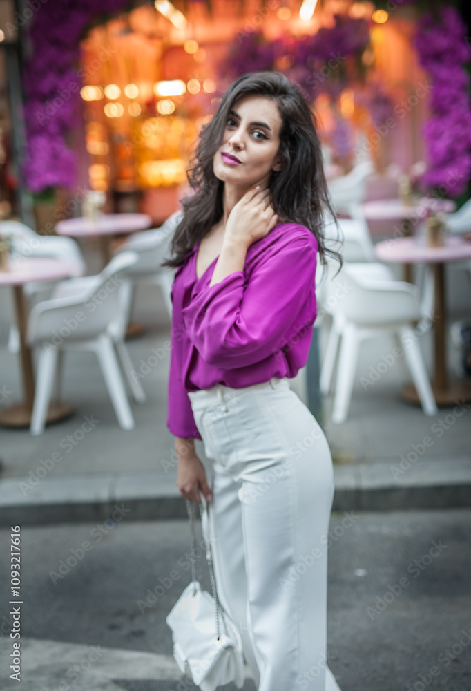 Young beautiful brunette woman in a pink jacket and white pants posing on the street. Portrait of a beautiful young woman in a purple blouse and white pants posing in an outdoor cafe on the street