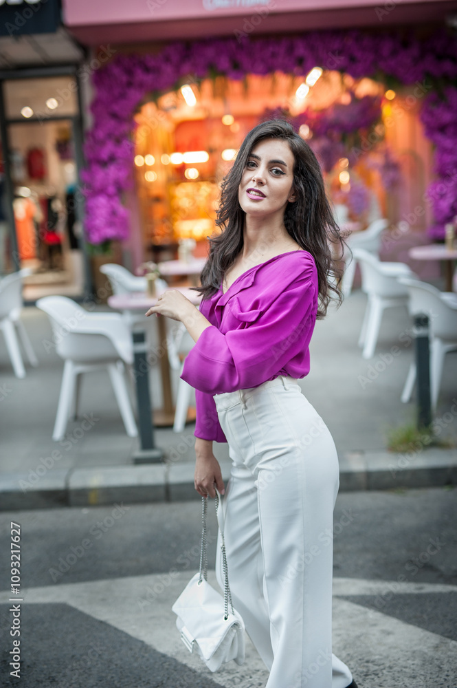 Young beautiful brunette woman in a pink jacket and white pants posing on the street. Portrait of a beautiful young woman in a purple blouse and white pants posing in an outdoor cafe on the street