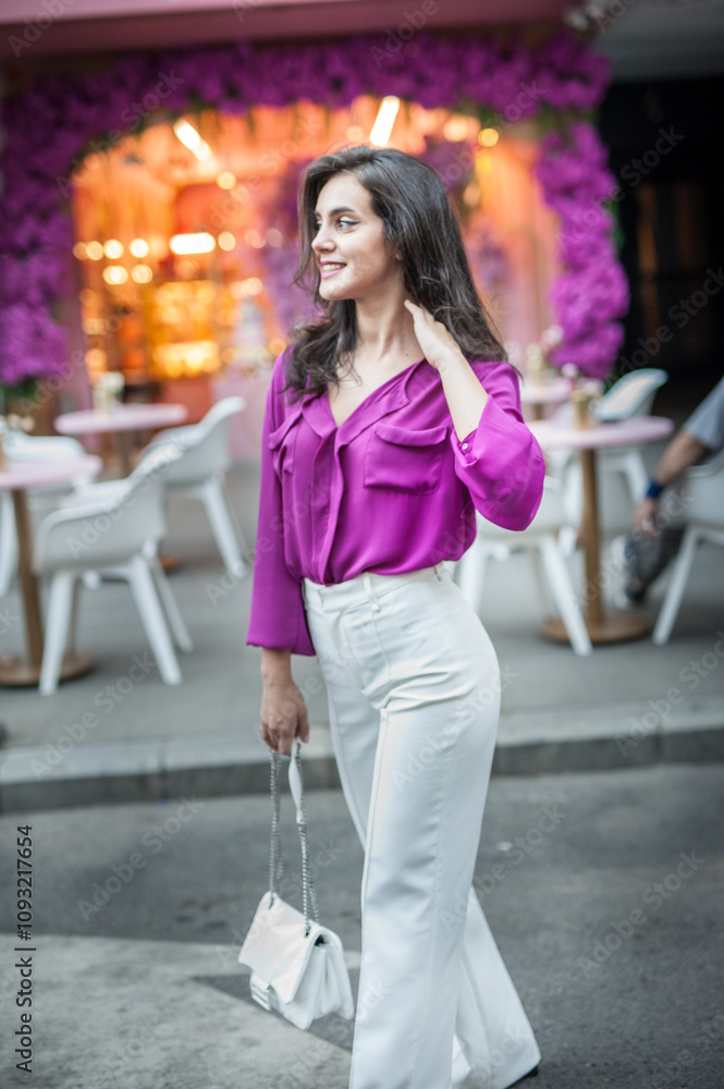 Young beautiful brunette woman in a pink jacket and white pants posing on the street. Portrait of a beautiful young woman in a purple blouse and white pants posing in an outdoor cafe on the street