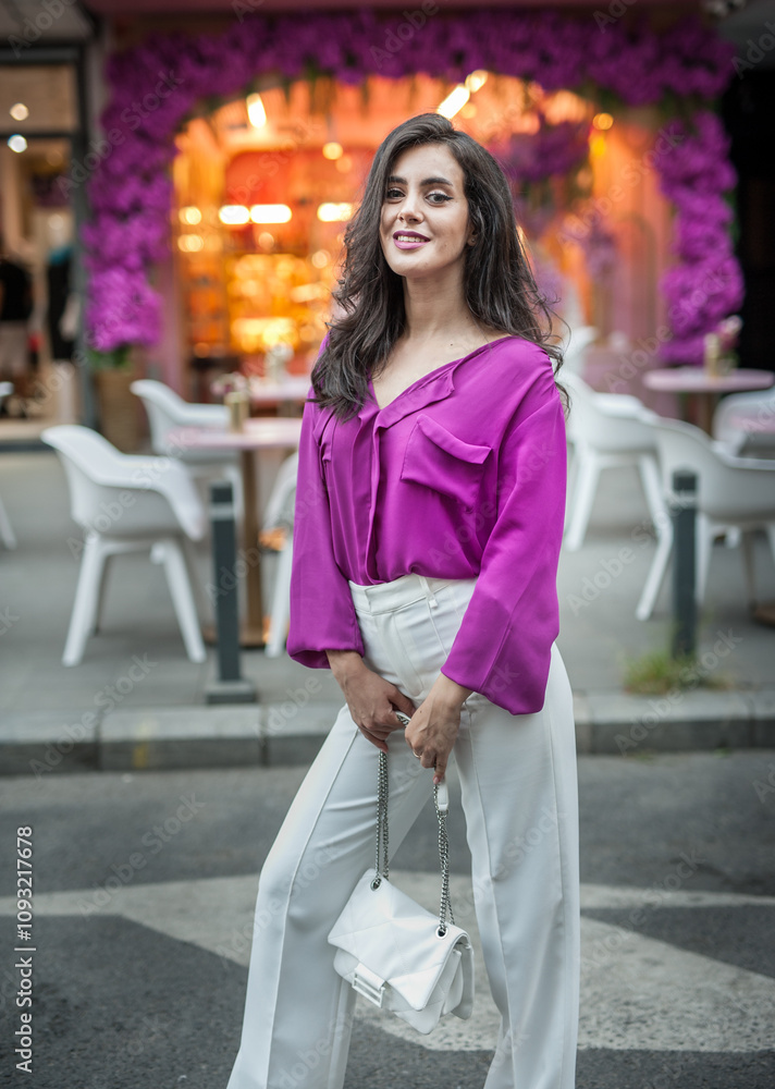 Young beautiful brunette woman in a pink jacket and white pants posing on the street. Portrait of a beautiful young woman in a purple blouse and white pants posing in an outdoor cafe on the street