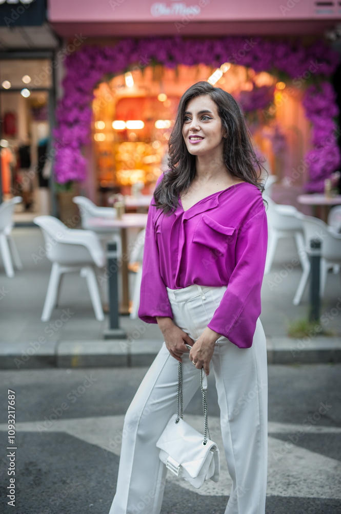 Young beautiful brunette woman in a pink jacket and white pants posing on the street. Portrait of a beautiful young woman in a purple blouse and white pants posing in an outdoor cafe on the street