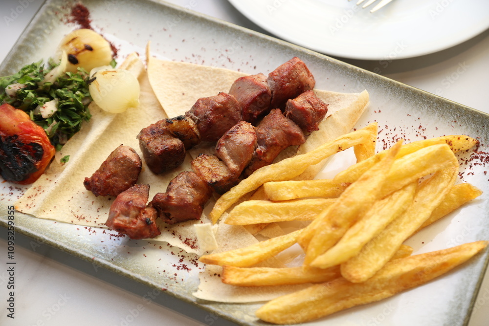 A Lebanese mixed grill platter featuring chargrilled beef kebabs, chicken skewers, and lamb skewers, served with French fries, garlic sauce, grilled tomatoes, fresh parsley, and sumac on flatbread, of