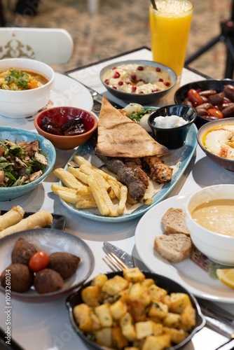 A vibrant Lebanese breakfast feast featuring mixed grill, hummus with pomegranate garnish, fattoush, lentil soup, kibbeh, fried potatoes, cheese rolls, garlic sauce, and fresh salad, served with flatb