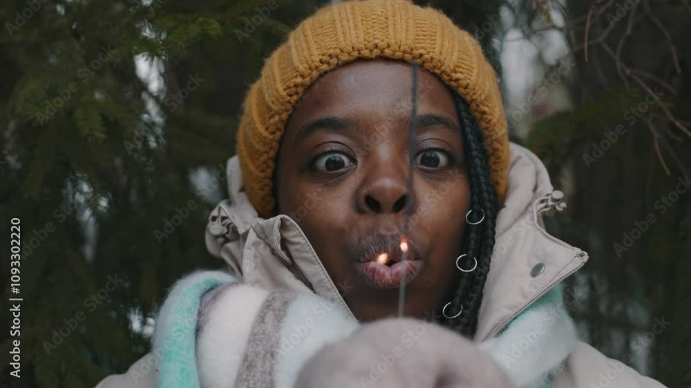 Close-up rack focus portrait of happy Black young woman with burning down sparkler, she blowing it out and laughing while standing under branches of fir tree in winter forest Close-up rack focus portrait of happy Black young woman with burning down sparkler, she blowing it out and laughing while standing under branches of fir tree in winter forest