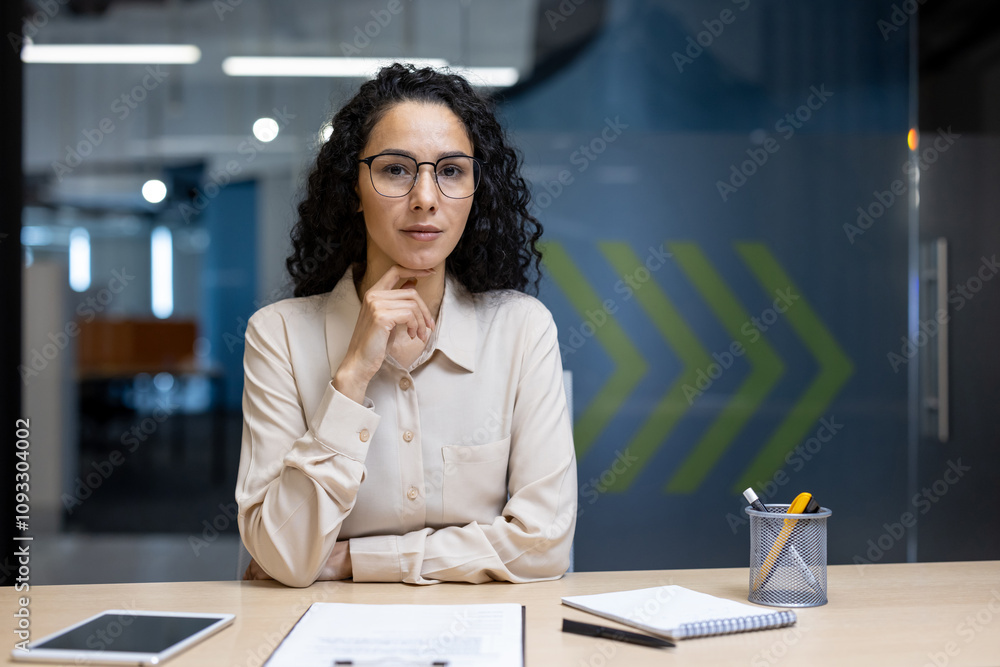 Hispanic businesswoman sitting confidently at office desk with notepad and tablet. Professional demeanor suggests leadership and determination in corporate setting. Hispanic businesswoman sitting confidently at office desk with notepad and tablet. Professional demeanor suggests leadership and determination in corporate setting.