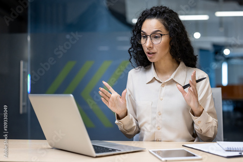 Hispanic business woman in office communicates actively during online meeting. Engaging in remote work, she uses laptop with notebook and phone by side, exuding professionalism and concentration.