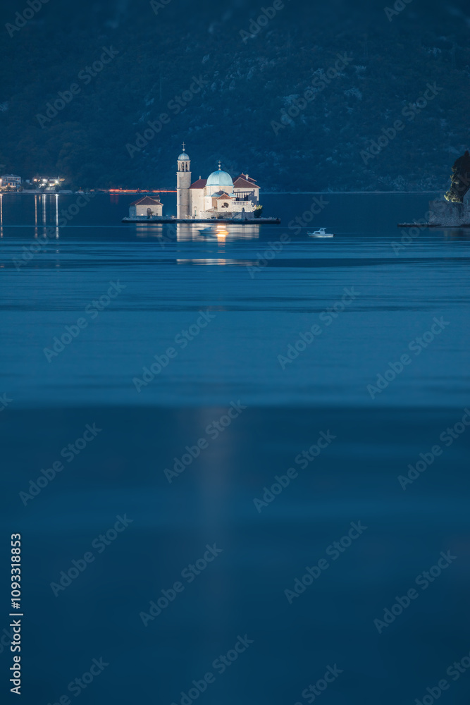 Fototapeta premium Scenic view of Our lady of the rocks island church glowing in the bay of Kotor, Montenegro at twilight with mountains in the background