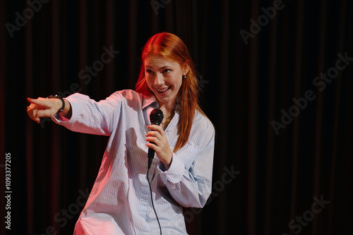 Fotografie Young Caucasian woman passionately performing stand-up comedy on stage, holding microphone and pointing toward audience