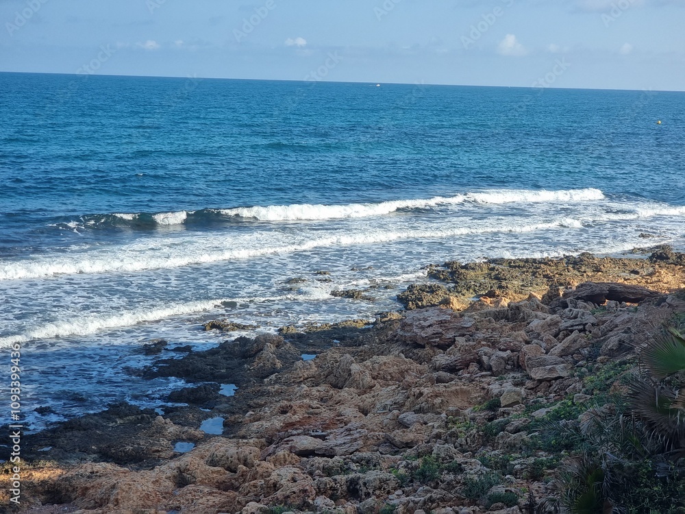 Szene am Meer mit markanten Steine im Vordergrund, die von sanften Wellen umspült werden. Am Rand wachsen vereinzelt grüne Pflanzen, während der Horizont unter einem klaren Himmel in der Ferne verläuf Szene am Meer mit markanten Steine im Vordergrund, die von sanften Wellen umspült werden. Am Rand wachsen vereinzelt grüne Pflanzen, während der Horizont unter einem klaren Himmel in der Ferne verläuf